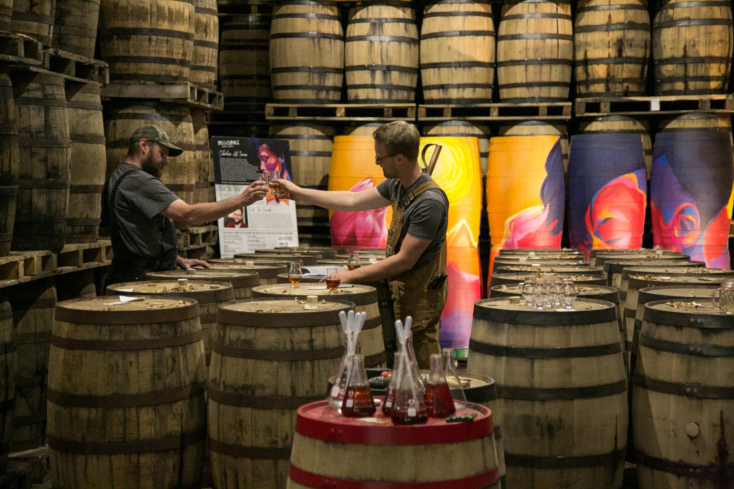 Two men drinking whiskey in room full of barrels